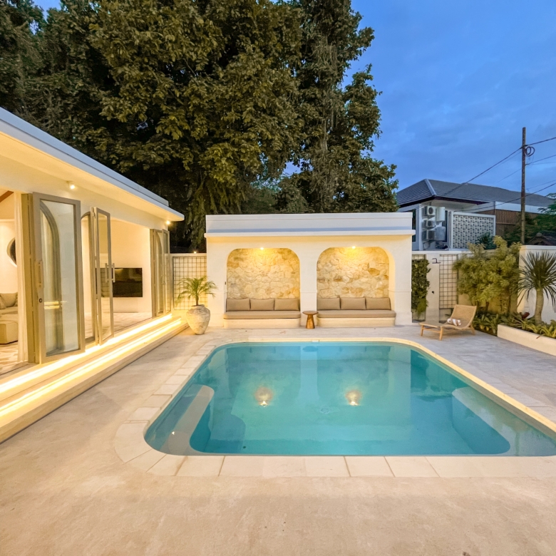 Contemporary backyard with an in-ground pool, white stone patio, and illuminated steps leading to a covered seating area at dusk.