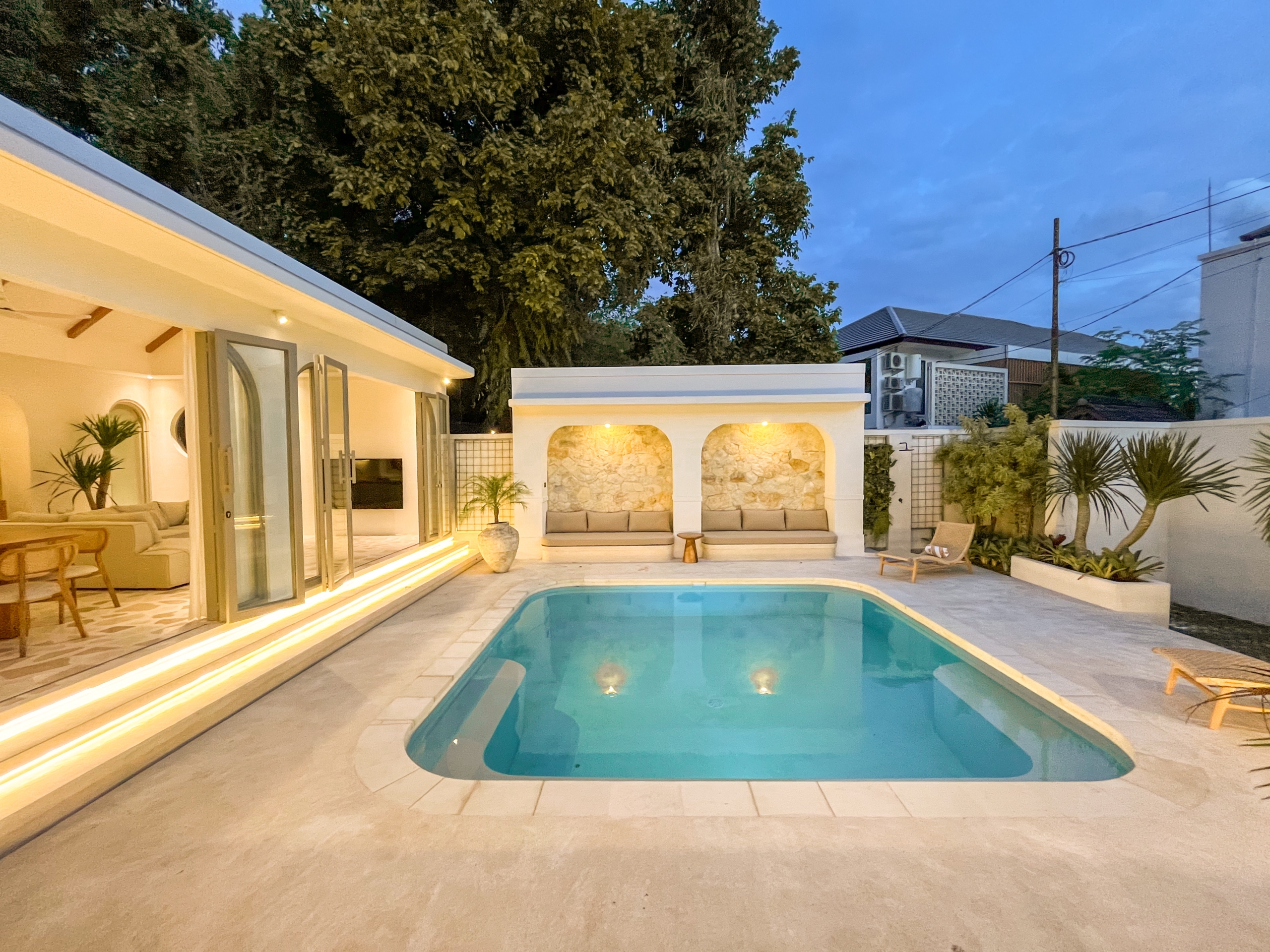 IMG_0599_4633-6a38c7b3bc242 Contemporary backyard with an in-ground pool, white stone patio, and illuminated steps leading to a covered seating area at dusk.