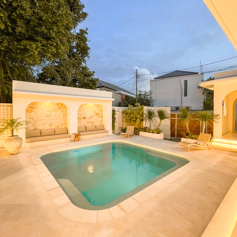Backyard pool area with built‑in lounge seating, white arches, and warm lighting at dusk.