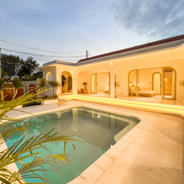 Modern white villa terrace at dusk with arched openings, warm illumination, and a rectangular pool bordered by palm plants.