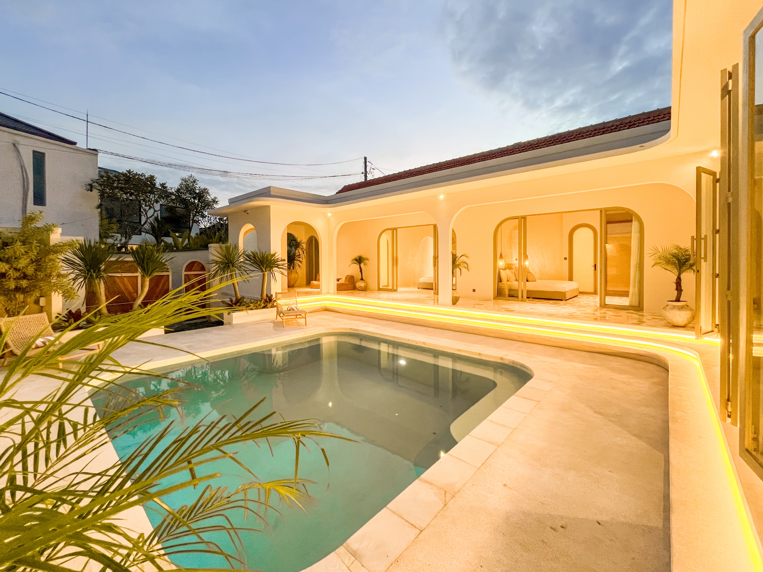 IMG_0602_4651-6a38c7b42c5c5 Modern white villa terrace at dusk with arched openings, warm illumination, and a rectangular pool bordered by palm plants.
