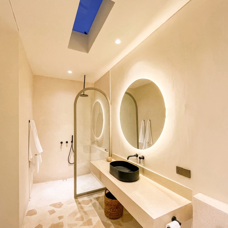 Spa-style bathroom showing double circular backlit mirrors above a beige countertop with a black vessel sink and woven stool, towels on a rack. (skylight visible)