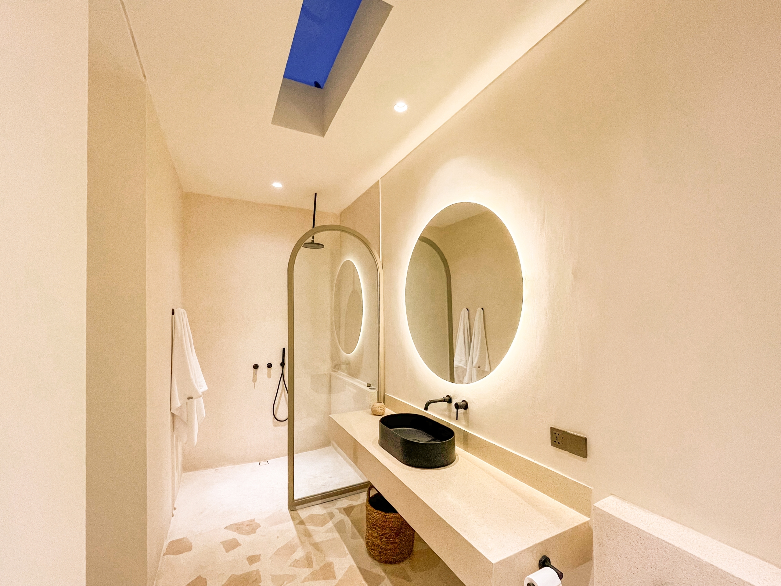 IMG_0636_4707-6a38c7b69a2c2 Spa-style bathroom showing double circular backlit mirrors above a beige countertop with a black vessel sink and woven stool, towels on a rack. (skylight visible)