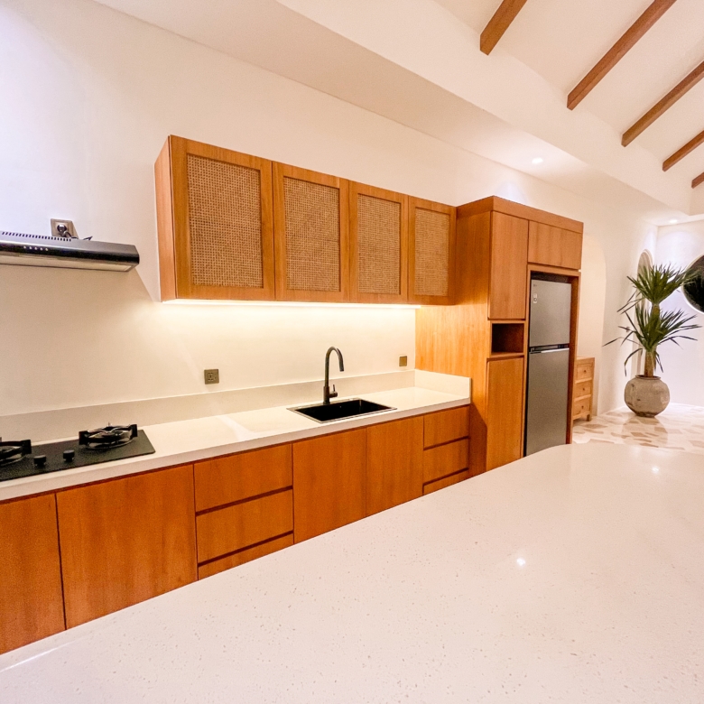 Modern kitchen with long white countertop, wooden cabinets, gas stove, and sink under a vaulted ceiling with wooden beams.