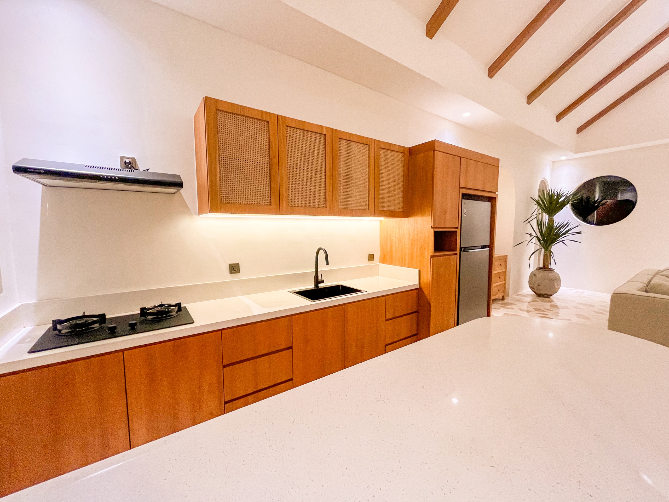 IMG_0654_5137-6a38c7b77ceca Modern kitchen with long white countertop, wooden cabinets, gas stove, and sink under a vaulted ceiling with wooden beams.