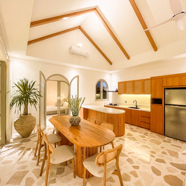 Open kitchen and dining area with vaulted ceiling, exposed wooden beams, a large wooden table, and a stainless fridge.