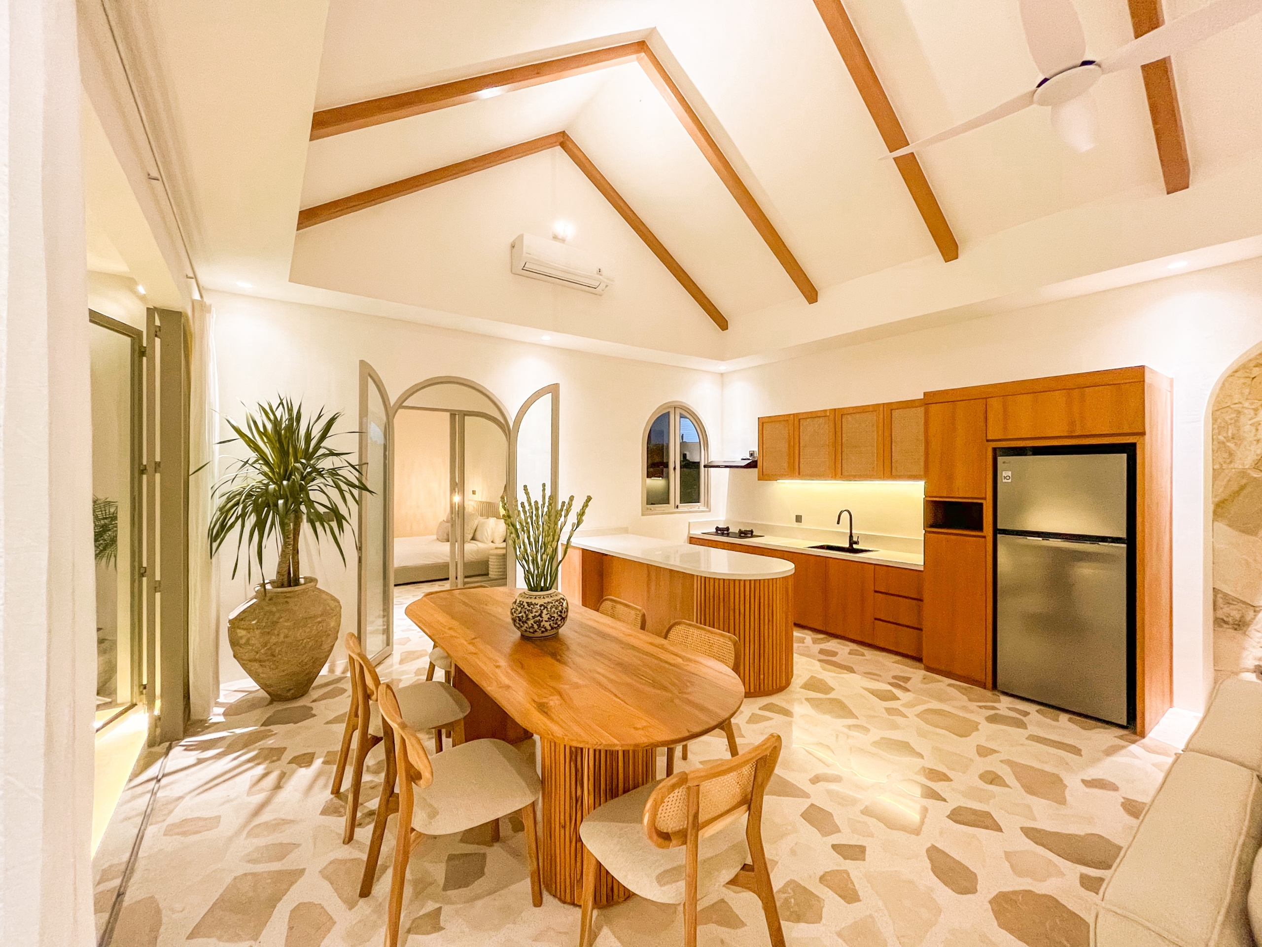 IMG_0664_4741-6a38c7b6ecd58 Open kitchen and dining area with vaulted ceiling, exposed wooden beams, a large wooden table, and a stainless fridge.