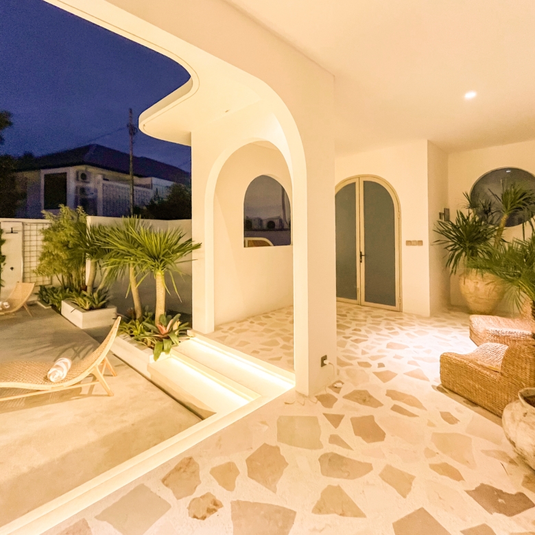 White arched patio with columns, wicker lounge chairs, and potted plants on a tiled floor at dusk/leisure lighting.