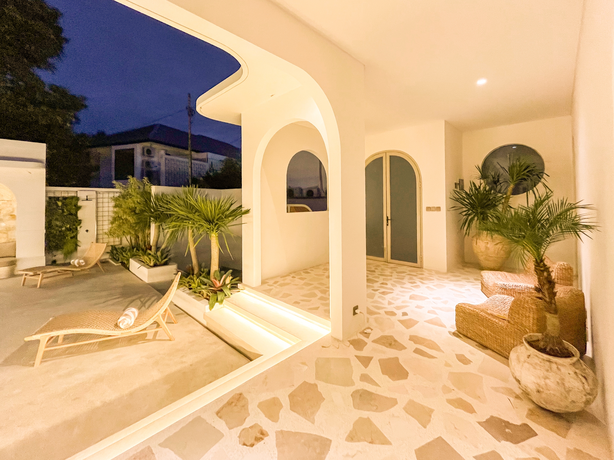 IMG_0679_4644-6a38c7b488424 White arched patio with columns, wicker lounge chairs, and potted plants on a tiled floor at dusk/leisure lighting.