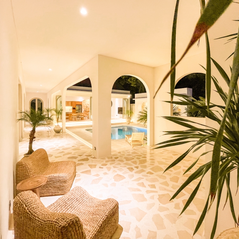 Indoor hotel atrium with arched openings, wicker seating, and a small pool framed by tropical plants under warm lighting.
