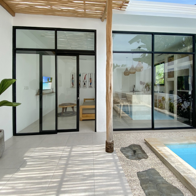 Modern glass sliding doors with black frames opening to a bright interior, white walls, and a poolside view outside.