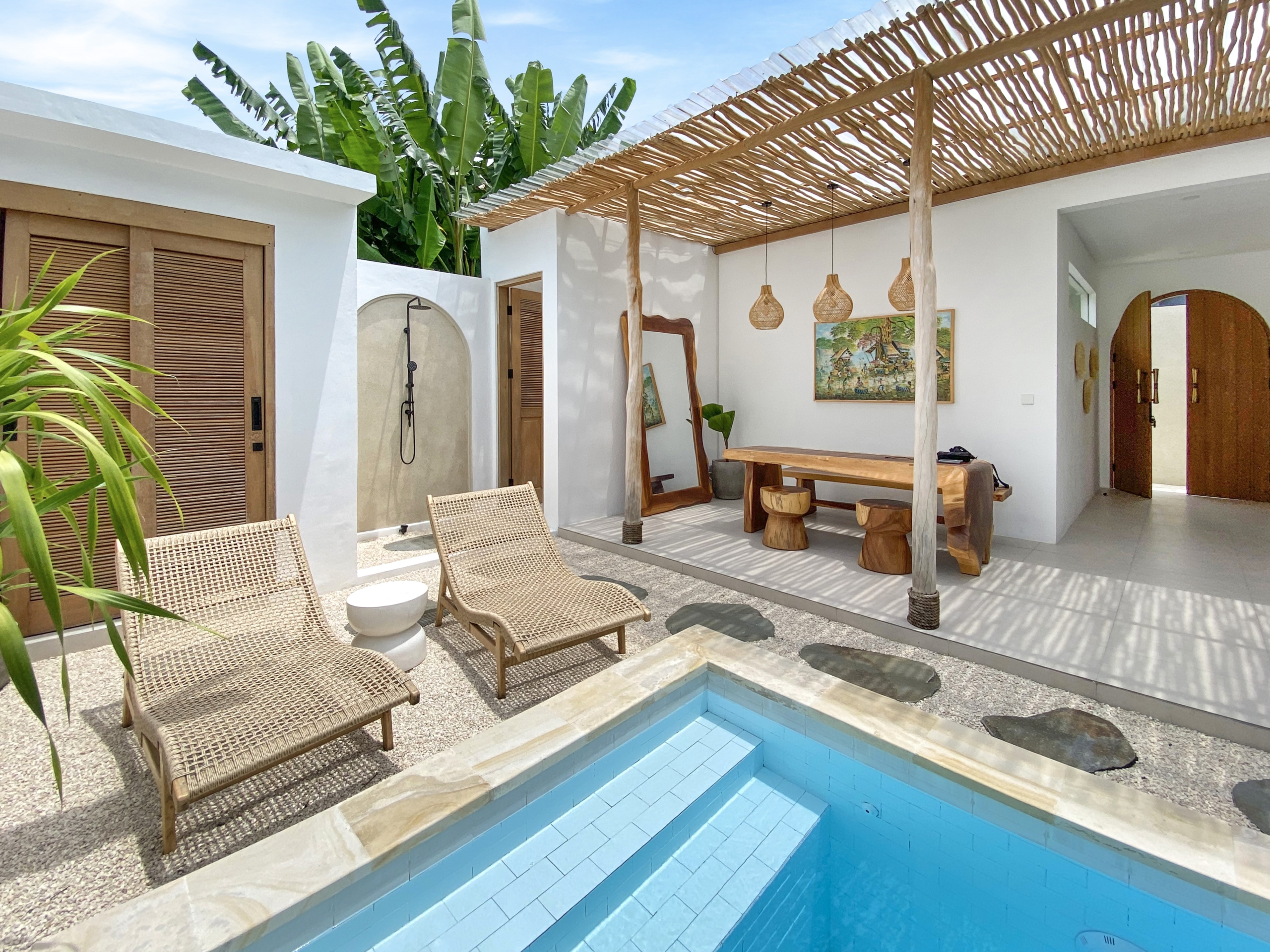 IMG_8109_5302-69ad009784f04 Outdoor patio with a blue tiled pool, wicker lounge chairs, and a wooden pergola over a white-walled seating area.
