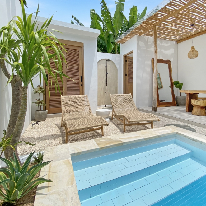 Outdoor pool area with light-blue tiled steps and a stone deck, wicker lounge chairs, and tropical plants against white walls and wooden doors.