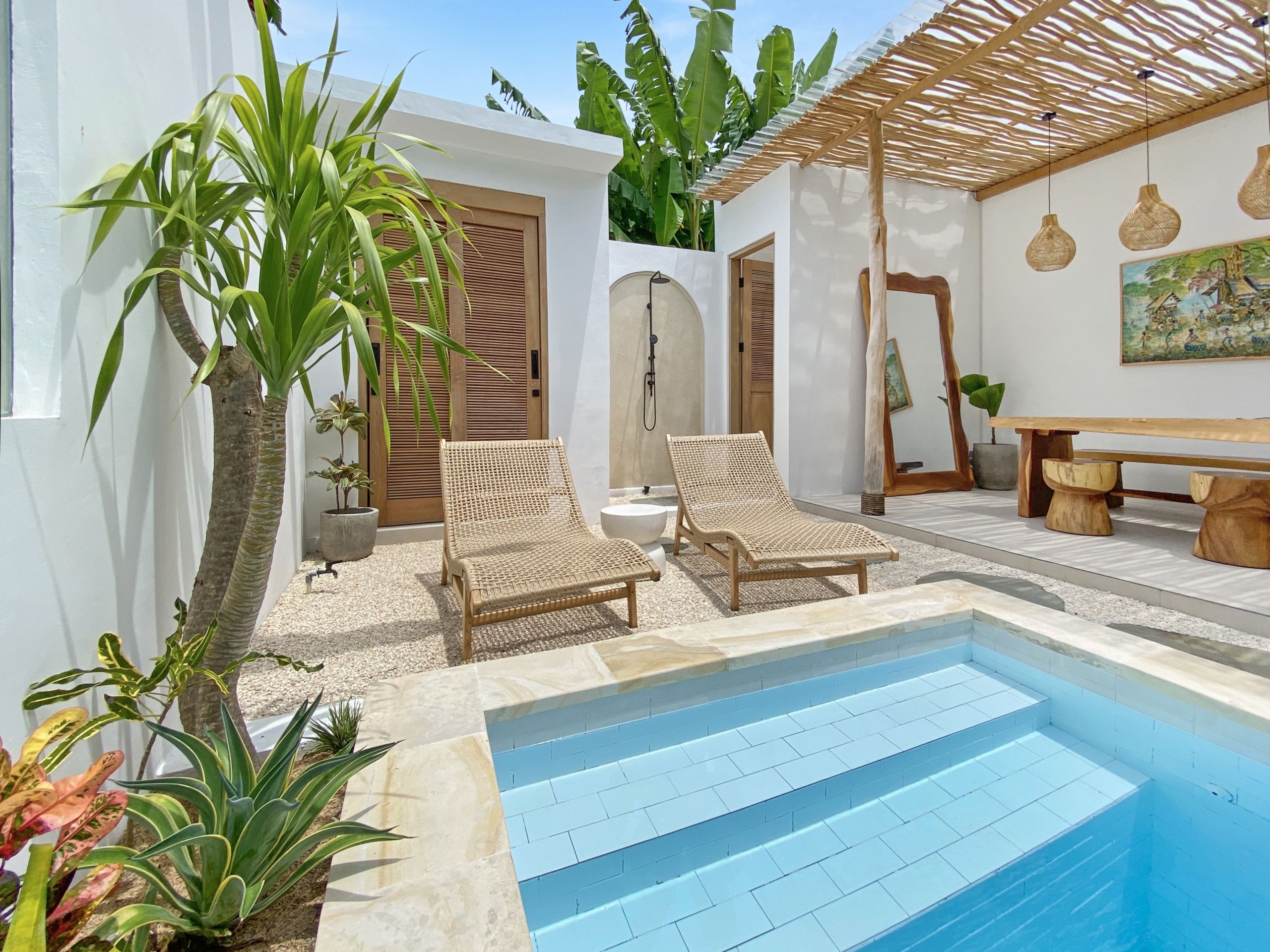 IMG_8110_5300-69ad0096bdc14 Outdoor pool area with light-blue tiled steps and a stone deck, wicker lounge chairs, and tropical plants against white walls and wooden doors.