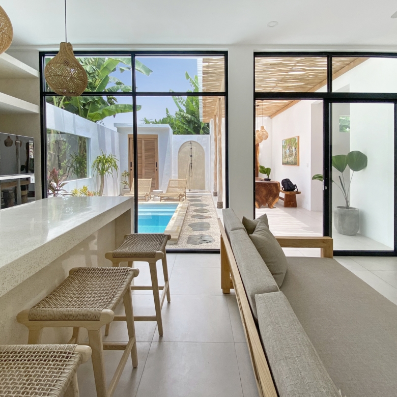 Open-plan living space with a white countertop bar, woven stools, and large glass doors showing a pool and tropical patio beyond.