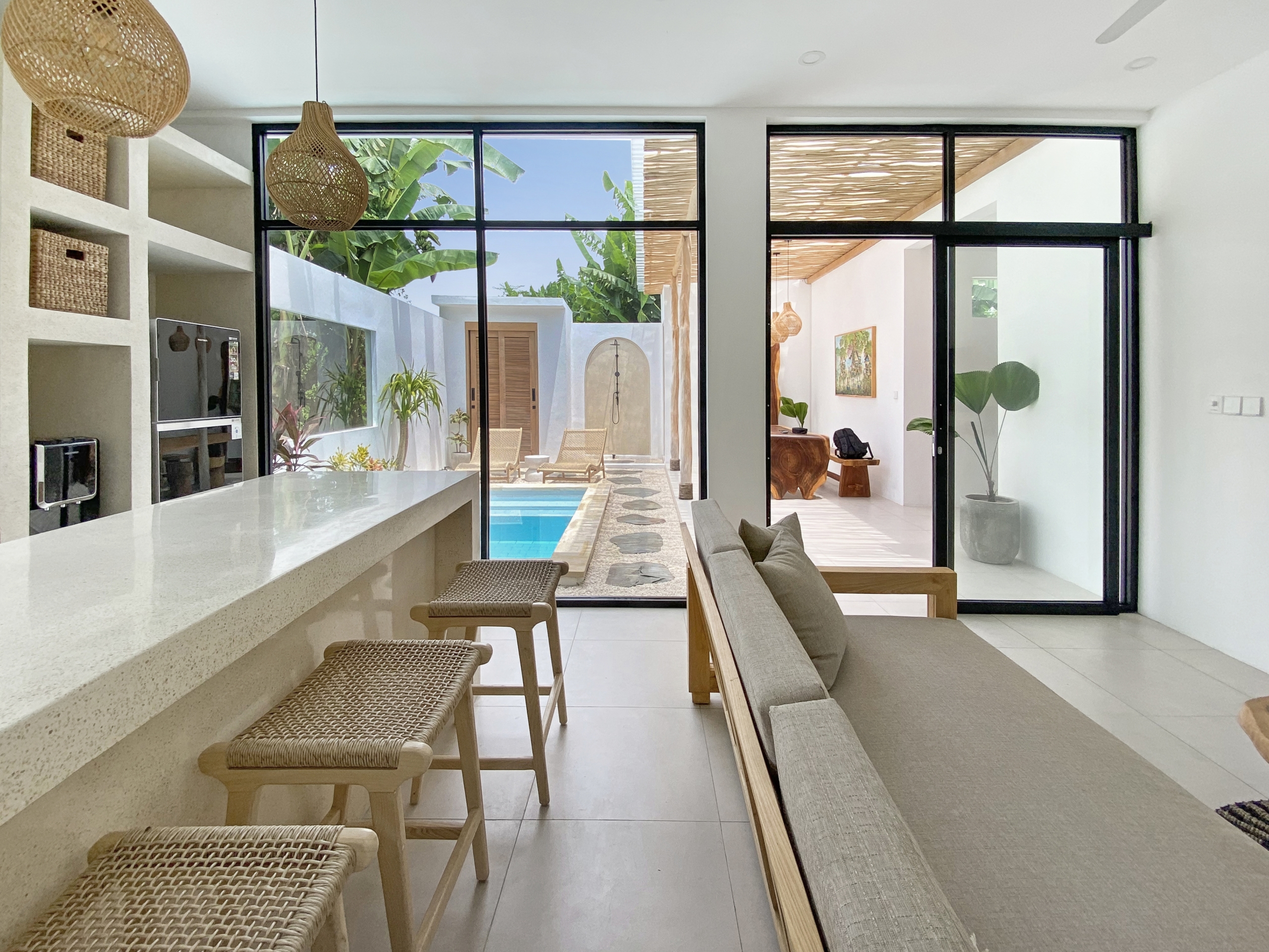 IMG_8115_5296-69ad00987df63 Open-plan living space with a white countertop bar, woven stools, and large glass doors showing a pool and tropical patio beyond.