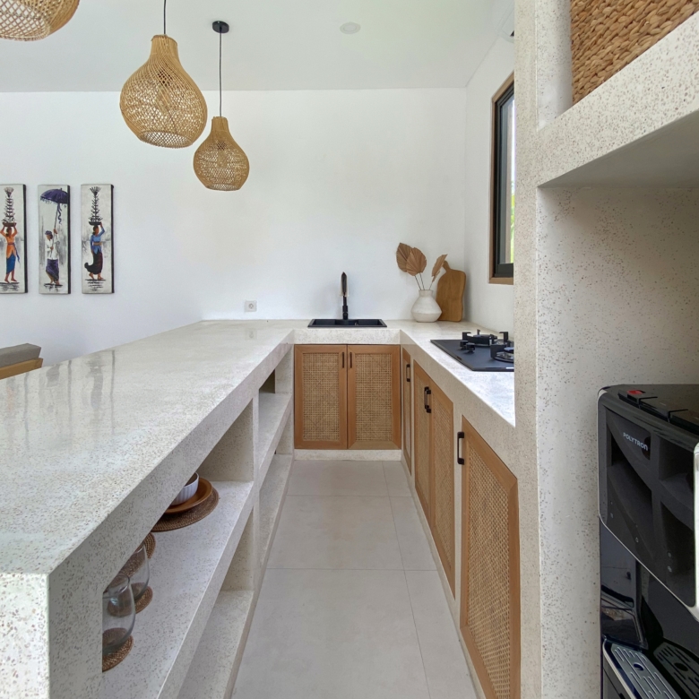 Modern kitchen with a long white terrazzo island, pendant wicker lights, and wooden cabinets beneath a white wall with framed art panels on the left.