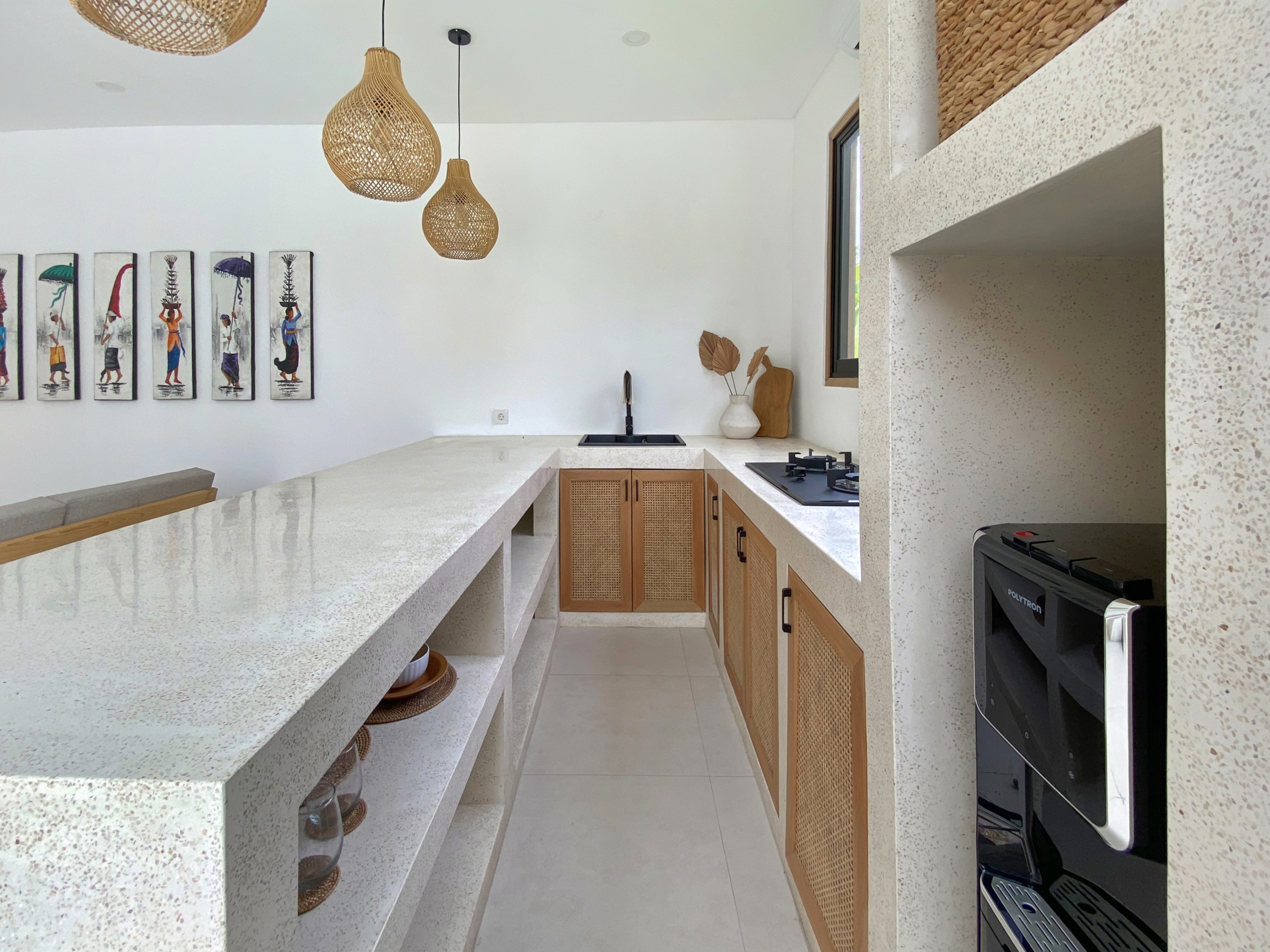 IMG_8117_5294-69ad0098a5c9d Modern kitchen with a long white terrazzo island, pendant wicker lights, and wooden cabinets beneath a white wall with framed art panels on the left.
