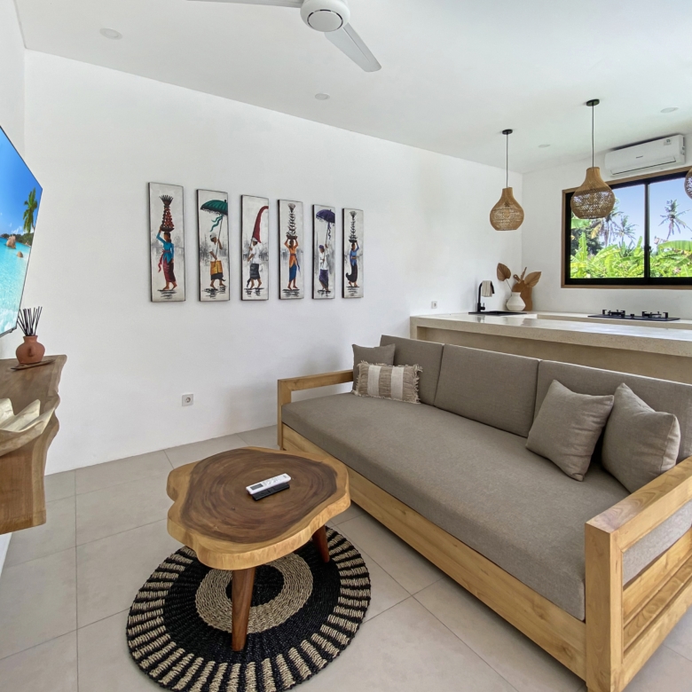 Living room with a beige sectional sofa, wooden coffee tables, and black-and-cream braided rug; tropical plants visible through large window. A row of framed art portraits on the wall.