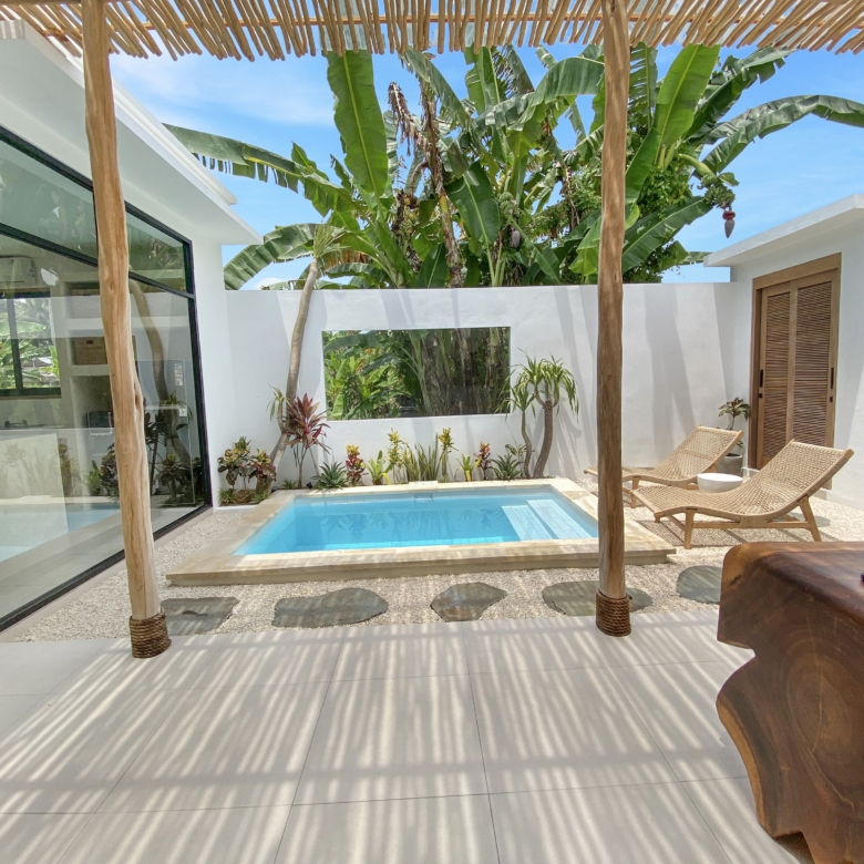 Poolside patio with a rectangular blue pool, white walls, glass doors, and wicker lounge chairs under a wooden shade structure surrounded by banana plants.