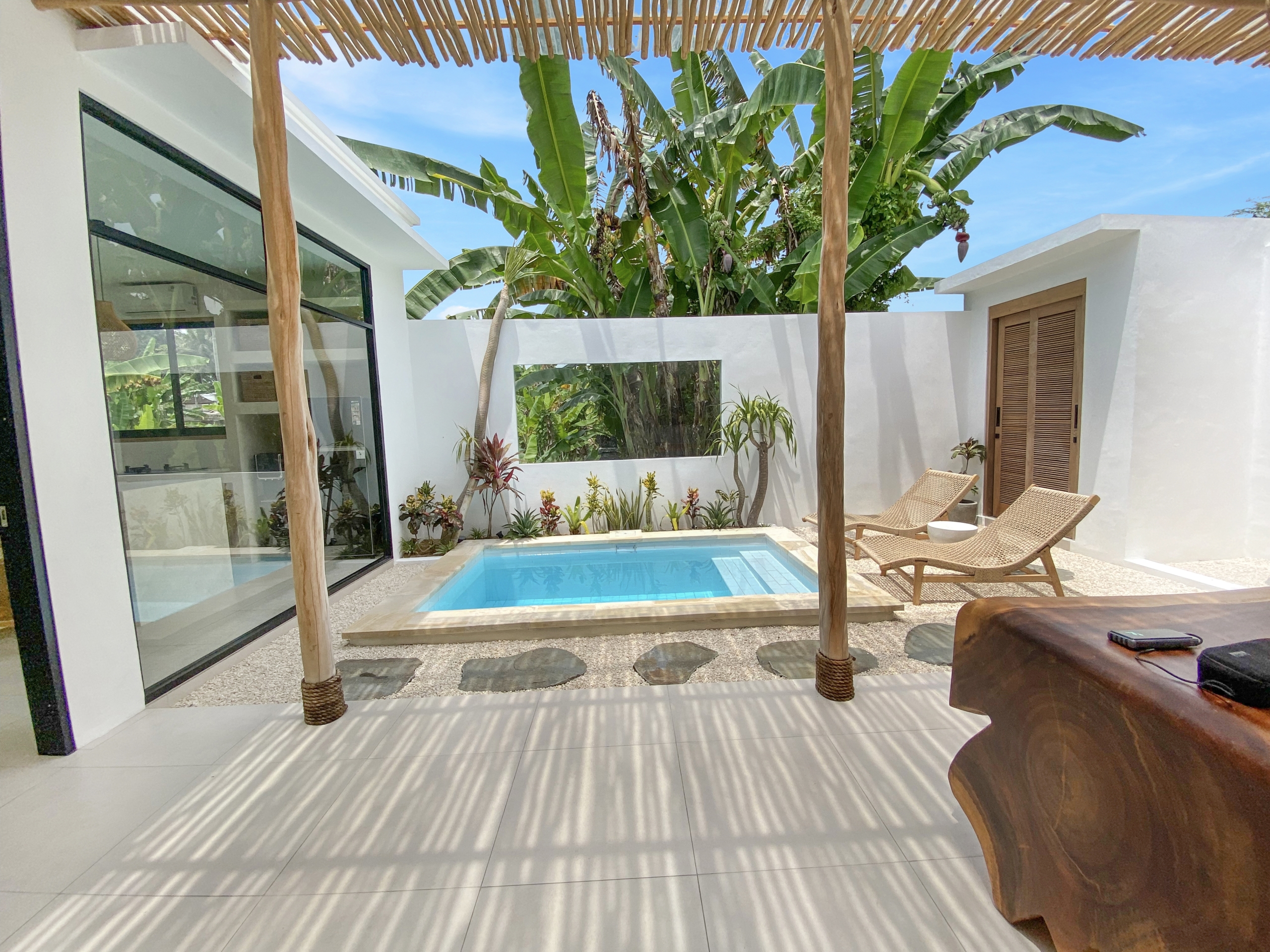 IMG_8120_5287-69ad0097a1d41 Poolside patio with a rectangular blue pool, white walls, glass doors, and wicker lounge chairs under a wooden shade structure surrounded by banana plants.
