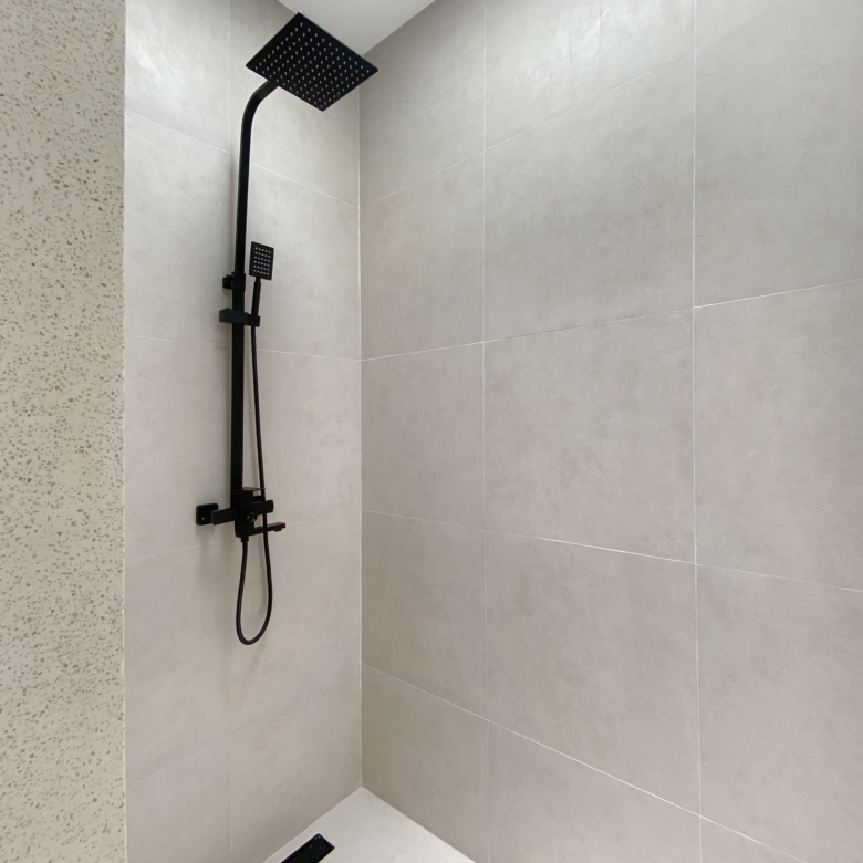 Matte black square rainfall showerhead with a handheld wand on a rail in a grey tiled shower area.