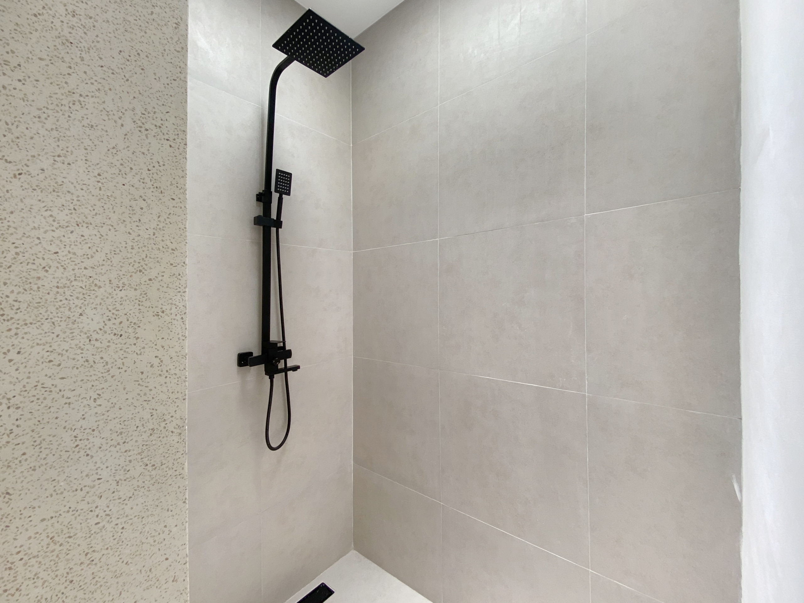 IMG_8129_5276-69ad00994f65d Matte black square rainfall showerhead with a handheld wand on a rail in a grey tiled shower area.