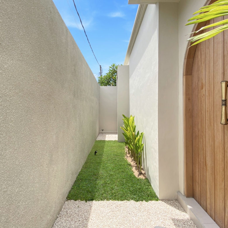 Narrow outdoor passage between a tall beige wall and a wooden gate, with a green lawn and tropical plants along the edge under a blue sky