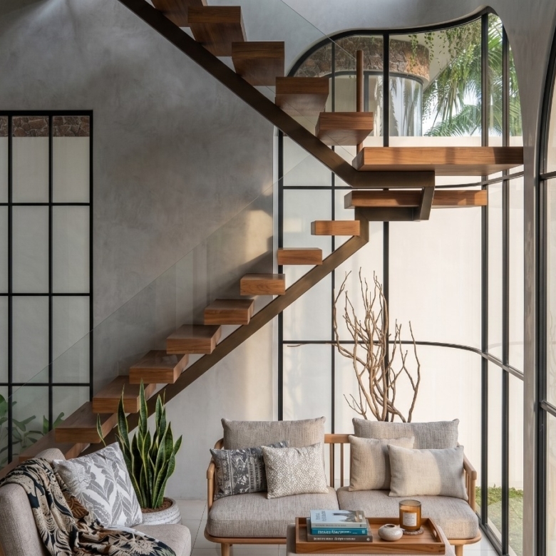 Sunlit living room with a floating wooden staircase along a glass wall, beige sofa, and potted plants outside the window.
