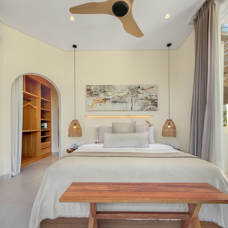 Bright bedroom interior with a large bed, two woven pendant lamps, and a wooden coffee table in the foreground.