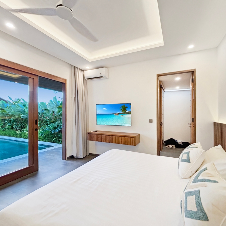 Hotel bedroom with white bedding, ceiling fan, wall TV, and sliding glass doors to a tropical pool and greenery outside.