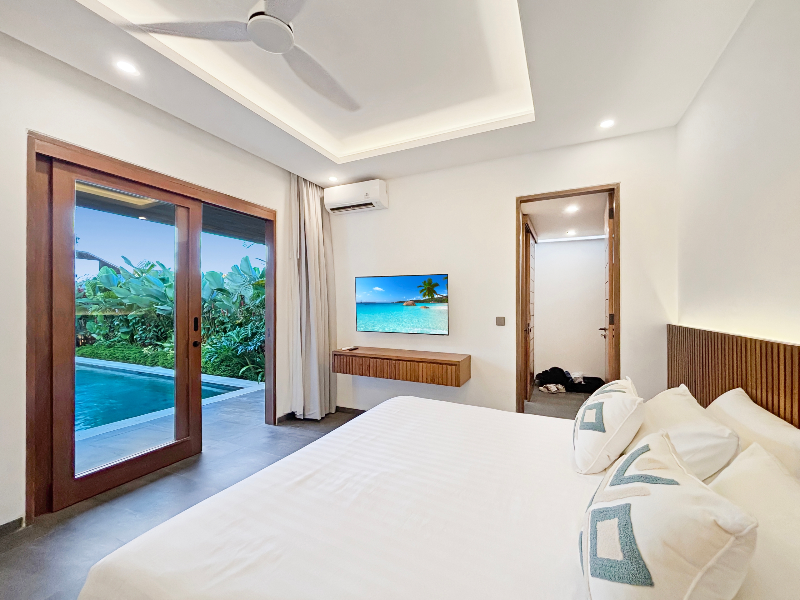 a29b8e8b000c4a122a8352fff053481e8e612af6 Hotel bedroom with white bedding, ceiling fan, wall TV, and sliding glass doors to a tropical pool and greenery outside.