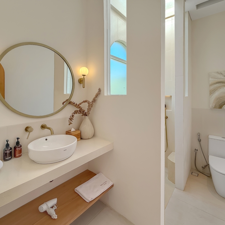 Modern bathroom with a round wall mirror, brass fixtures, and two globe wall sconces above a white countertop sink. A wooden shelf holds towels and a vase with dried stems; a window reflects blue sky nearby. A separate shower area and a toilet are visible to the right, with neutral-toned artwork on the wall.