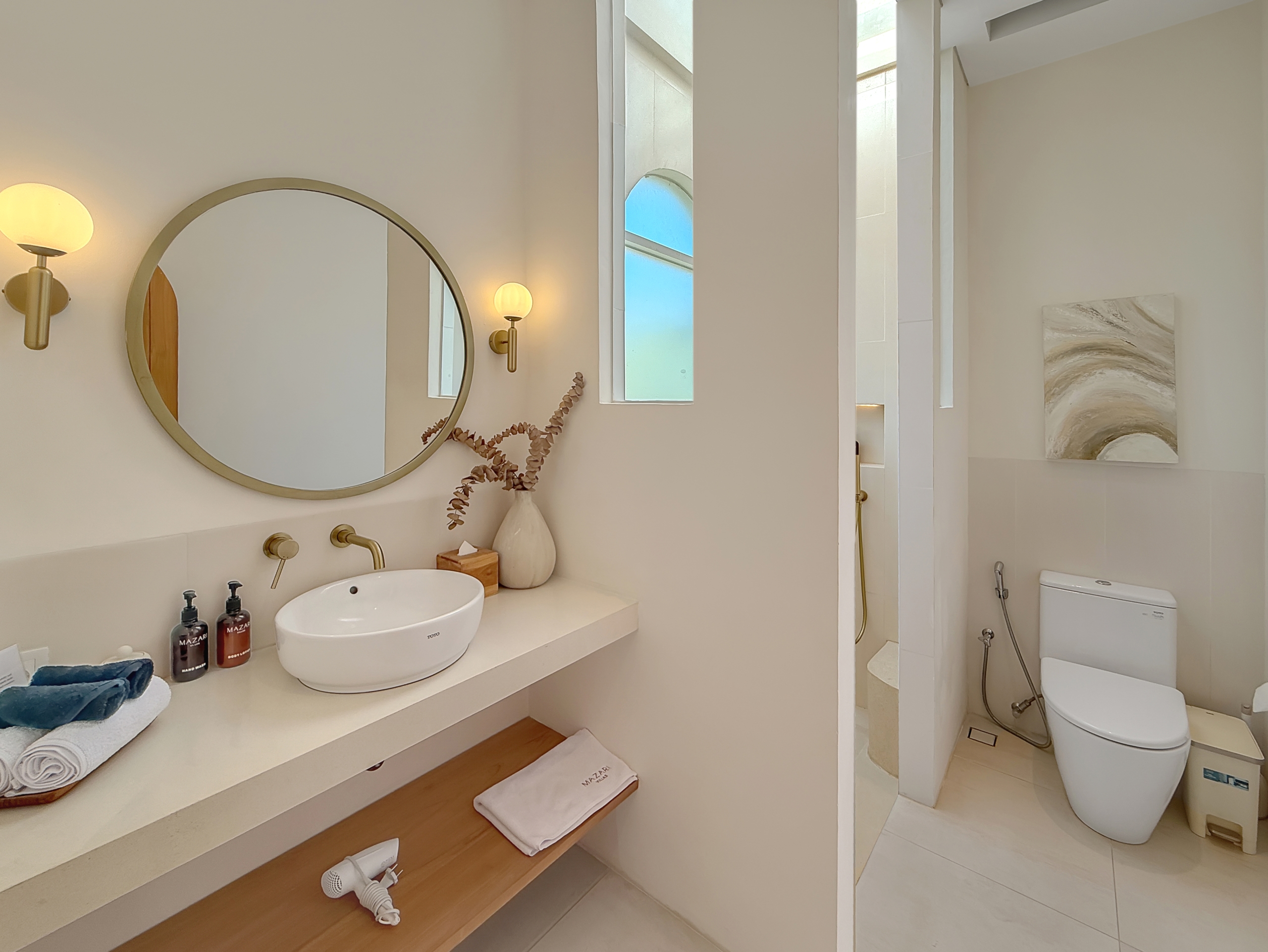 a2bfbaf7c71616c769f17389c1d539e91b0cbefe Modern bathroom with a round wall mirror, brass fixtures, and two globe wall sconces above a white countertop sink. A wooden shelf holds towels and a vase with dried stems; a window reflects blue sky nearby. A separate shower area and a toilet are visible to the right, with neutral-toned artwork on the wall.