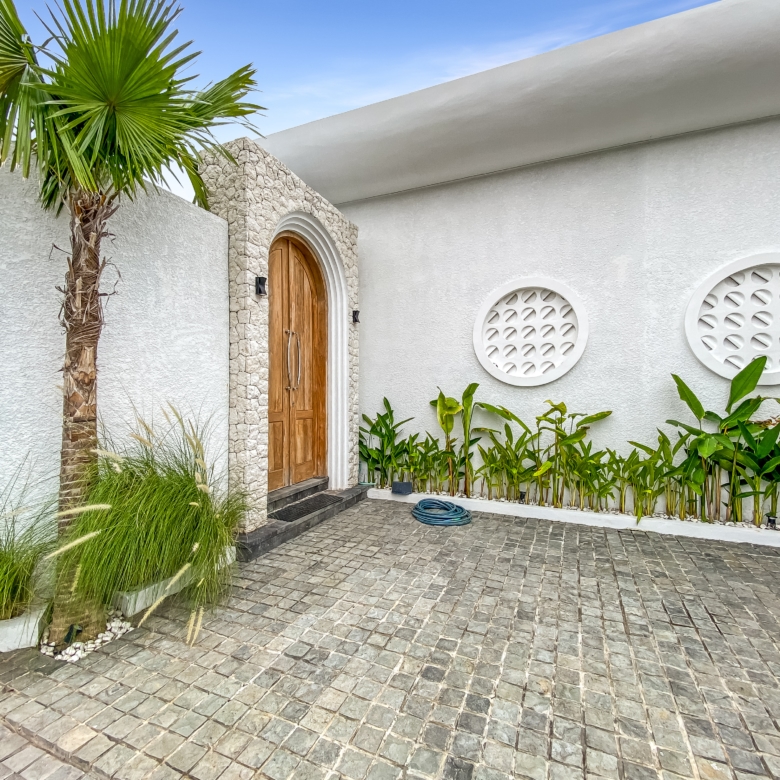 White exterior wall with a rounded stone-framed wooden door, palm tree, and decorative circular vents; tropical landscaping along the walkway.