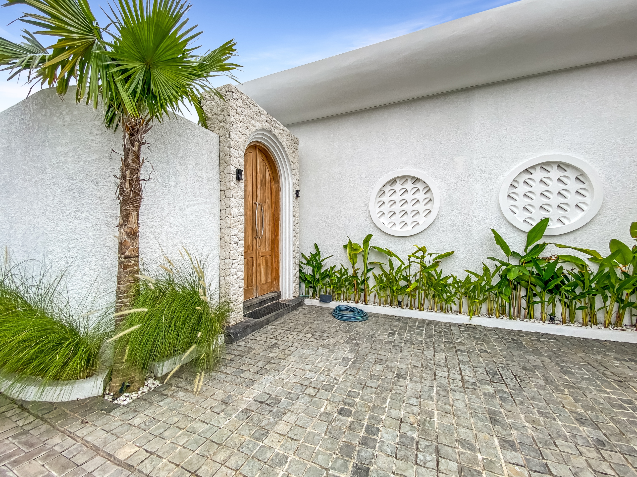 a464280a985dc407fa72ef710ea1a187b86d250d White exterior wall with a rounded stone-framed wooden door, palm tree, and decorative circular vents; tropical landscaping along the walkway.