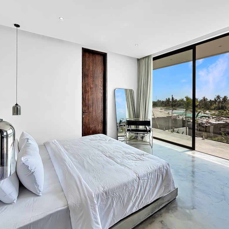 Bright modern bedroom with white bedding on a low platform bed, Nana glass wall showing palm trees and a blue sky outside.
