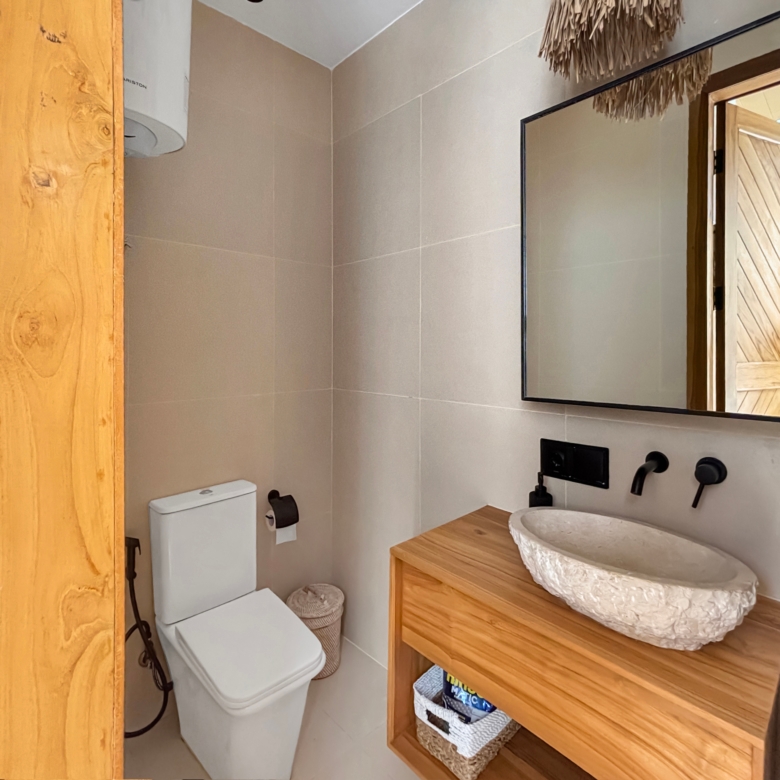 Bathroom with a white toilet and vessel sink on a wooden counter, reflected in a large wall mirror. The space has beige tile walls and a wooden door frame adjacent.