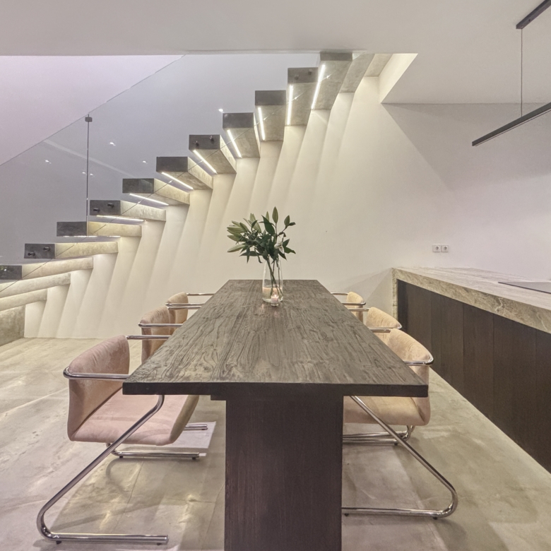Modern dining area with a long wooden table, beige chairs, and a glass wall beside a floating staircase on the left.