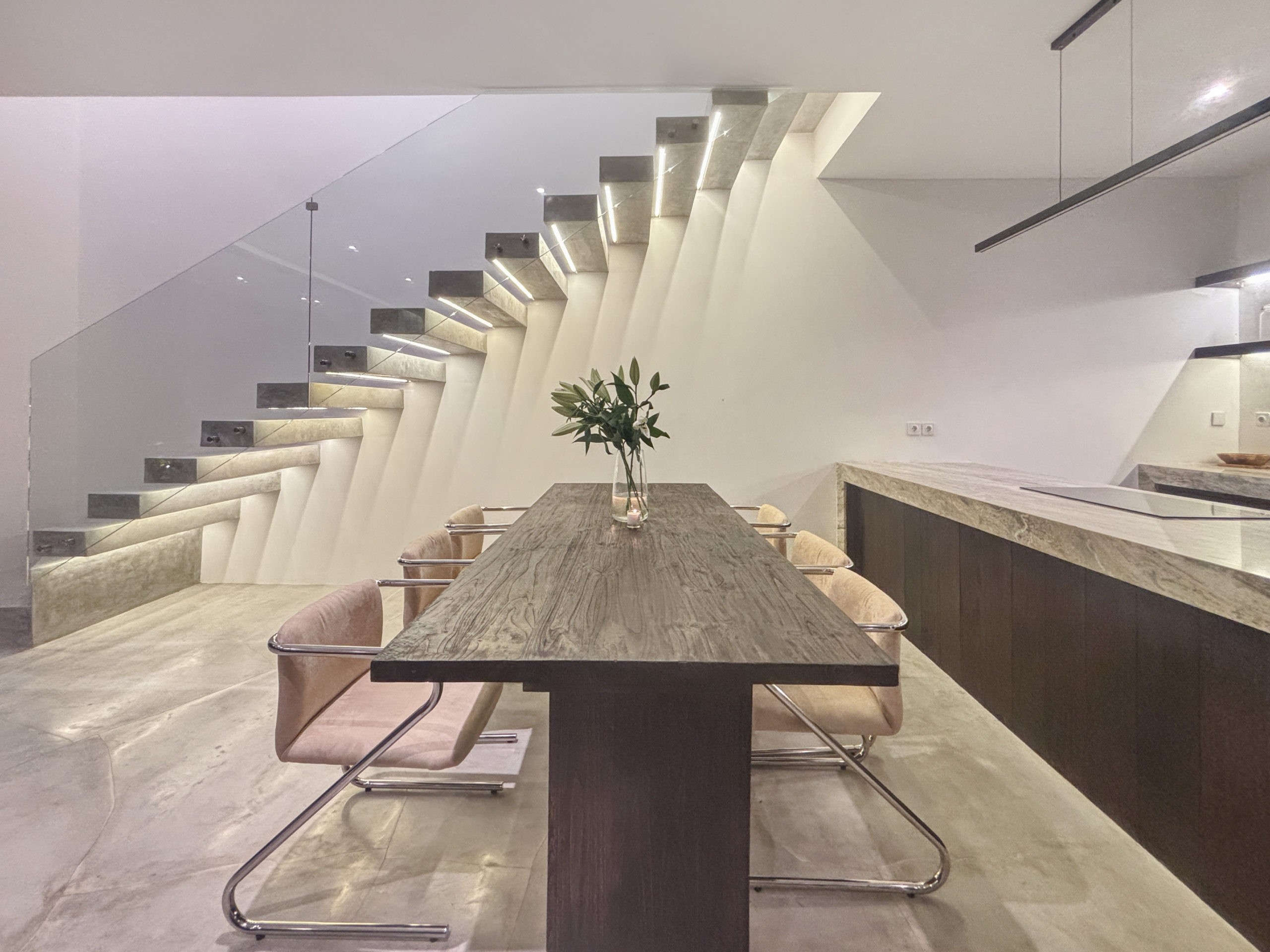 ab6ca28da248b07ca637f6490442bfa460f6a9f3_0942-6a05be06bf405 Modern dining area with a long wooden table, beige chairs, and a glass wall beside a floating staircase on the left.