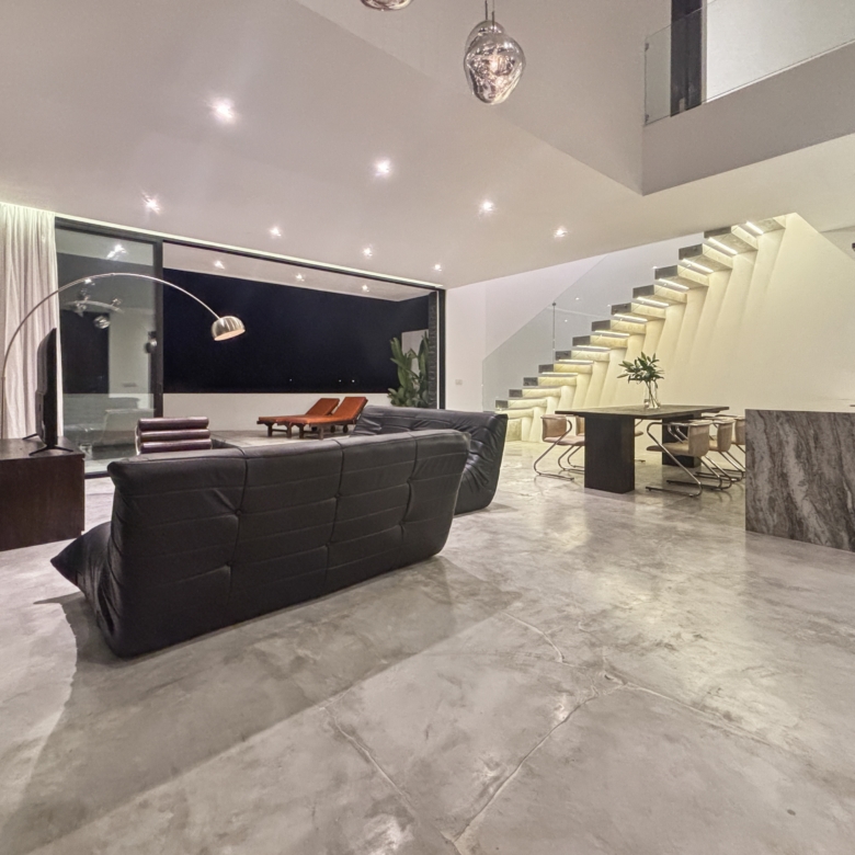 Open-plan modern living area with a black leather sofa, marble floors, and a glass-walled border around a night-lit interior space with a floating, illuminated staircase at right.