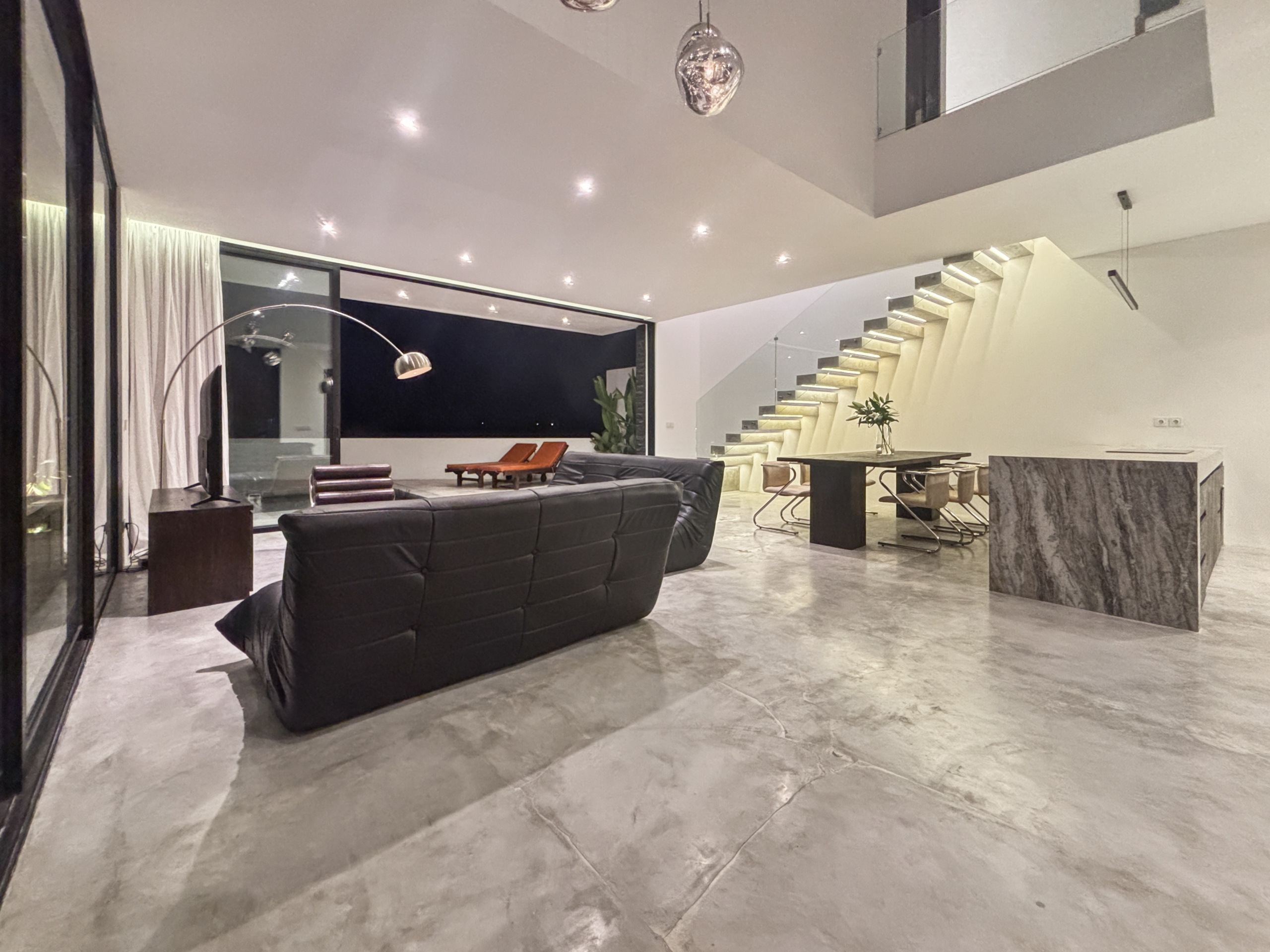 abdf6be059e05e1955d76fb508c21cfbd2a87dab_0870-6a05be05ac0c2 Open-plan modern living area with a black leather sofa, marble floors, and a glass-walled border around a night-lit interior space with a floating, illuminated staircase at right.