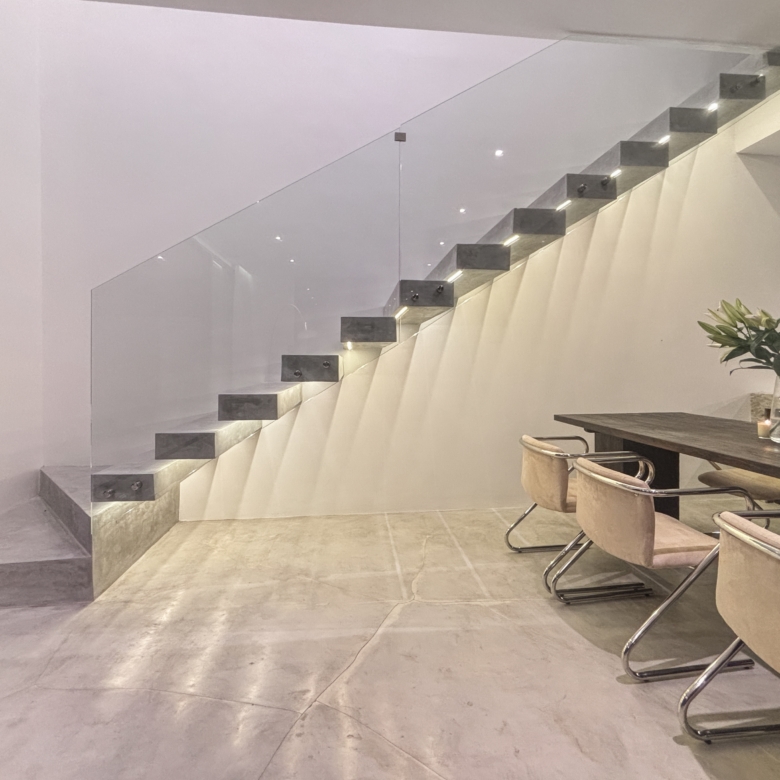 Modern interior with a glass-railed, floating staircase beside a dining table and beige chairs.