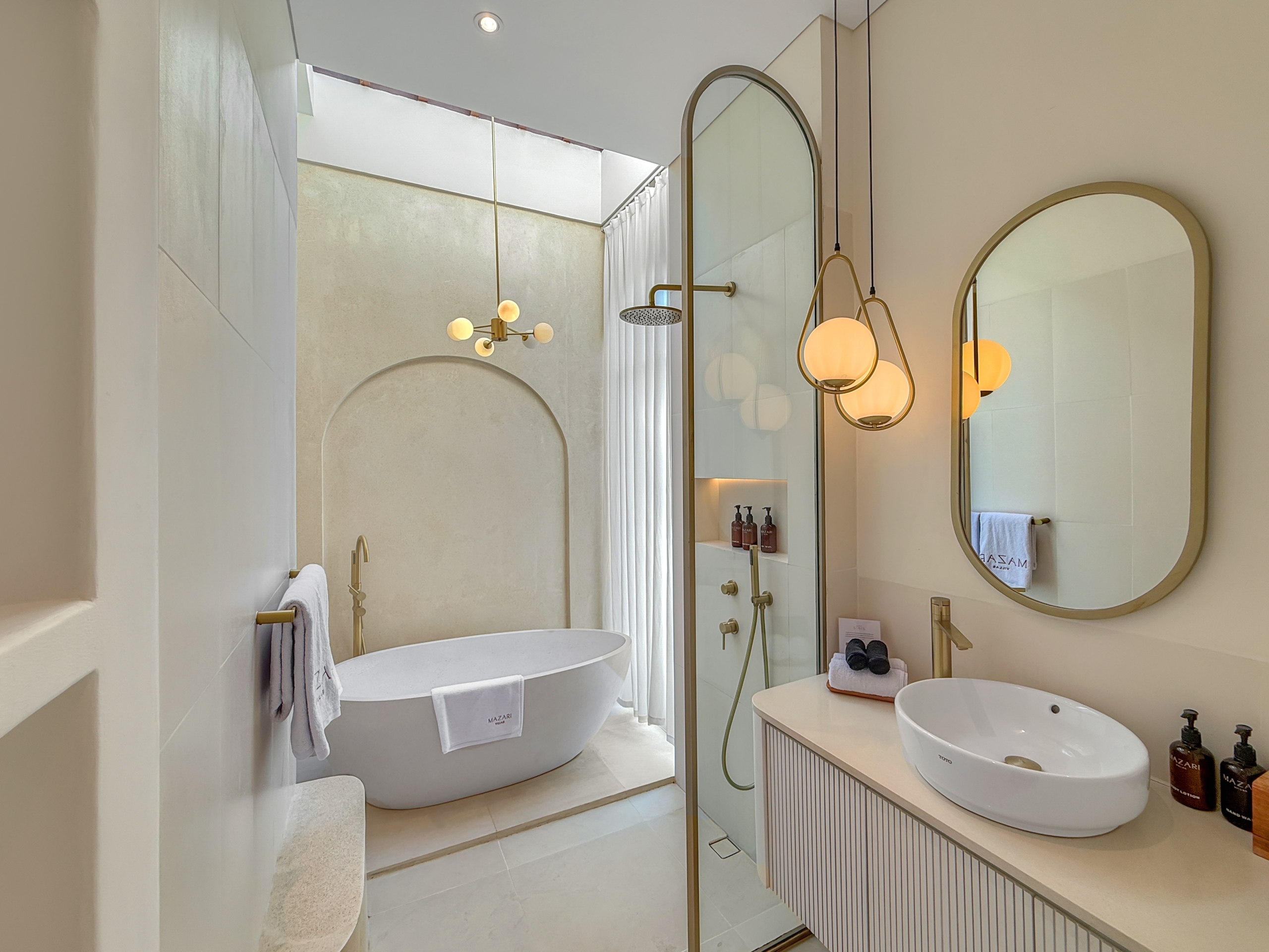 b16d51281113bb69e16d73260c076445e63b091e Luxurious bathroom with a white freestanding oval tub, glass shower, and gold fixtures beneath arched mirrors.