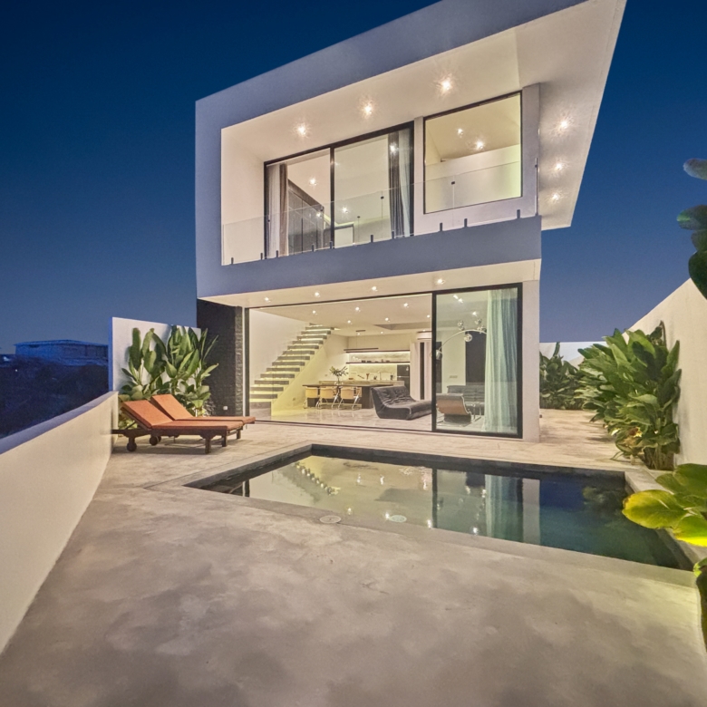 Night-time view of a modern white house with large glass walls, interior lighting visible, and a rectangular pool in the foreground.