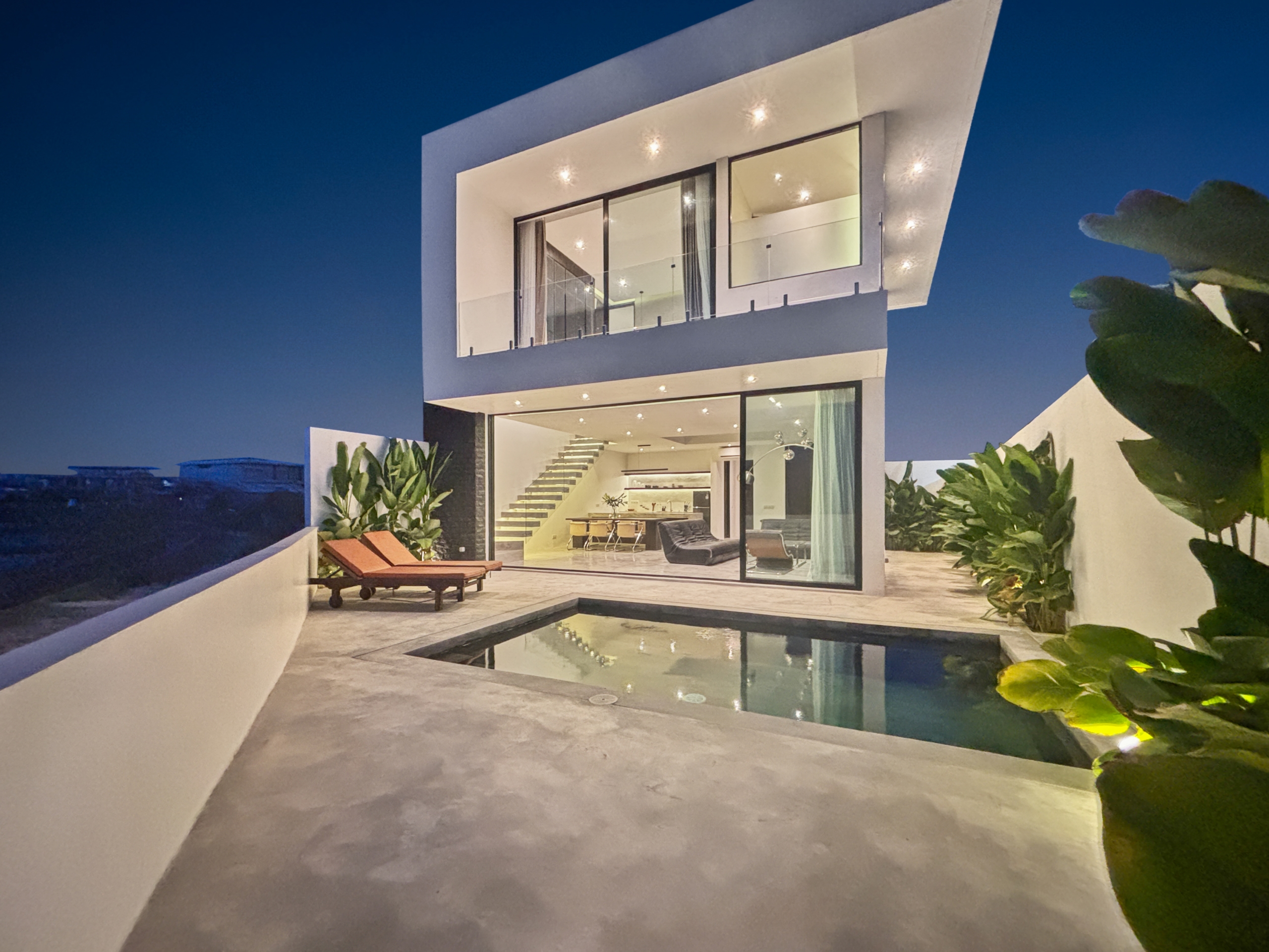 b1f5614a5b33f2f0aecb0c03f3002bf80ab63b19_0968-6a05be04628a5 Night-time view of a modern white house with large glass walls, interior lighting visible, and a rectangular pool in the foreground.