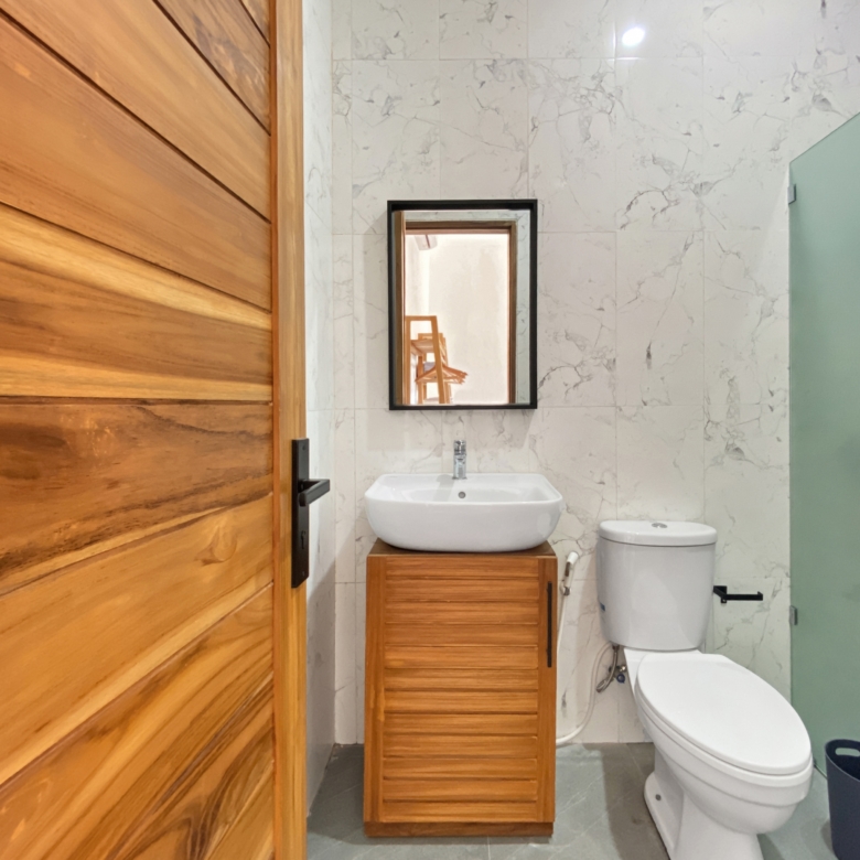 Wooden door on the left; white sink on a wooden cabinet, black-framed mirror, marble-tiled wall, and a toilet beside a frosted glass divider.
