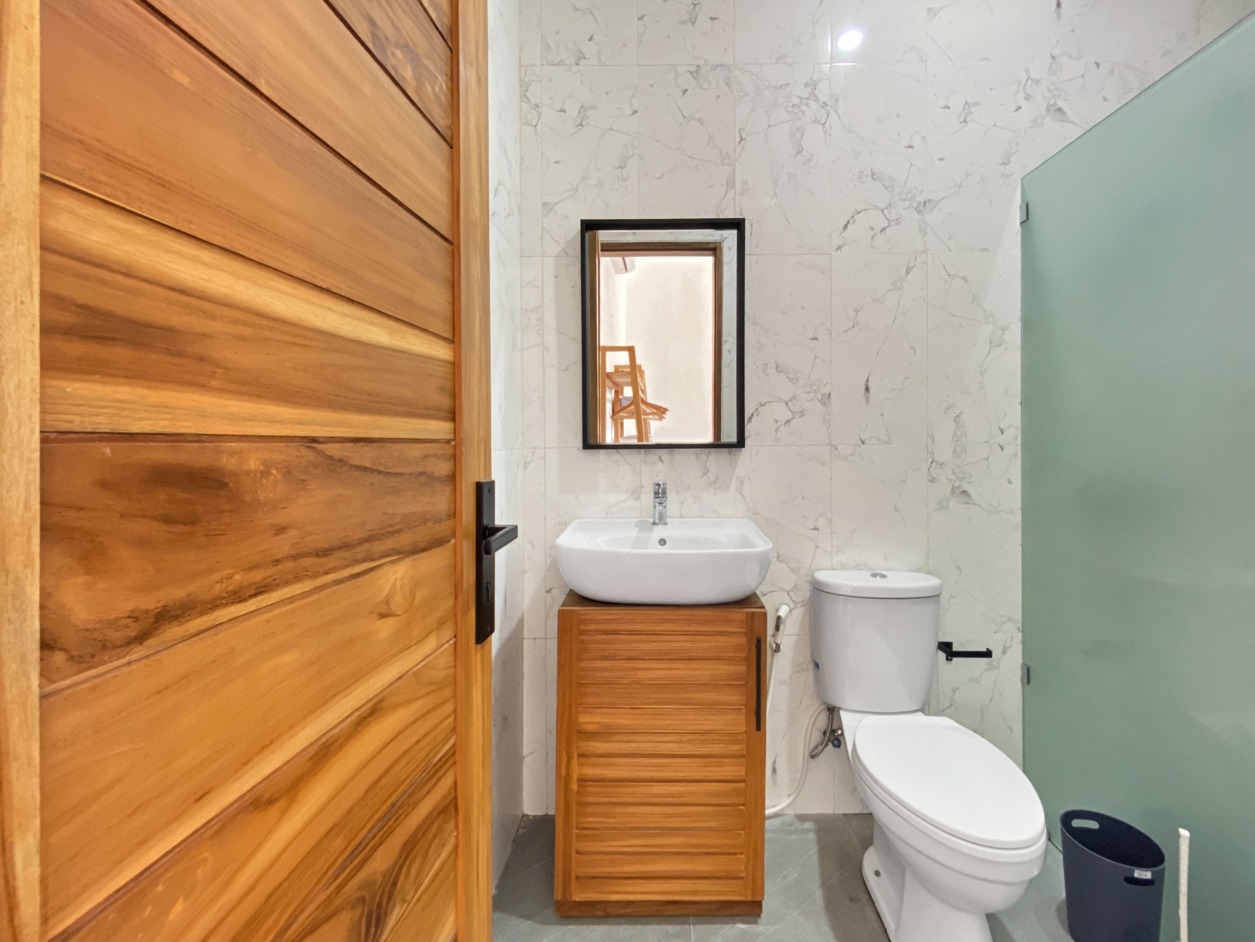 b679796a88b25eaa64cc9a82ac8b386d1afc055e Wooden door on the left; white sink on a wooden cabinet, black-framed mirror, marble-tiled wall, and a toilet beside a frosted glass divider.