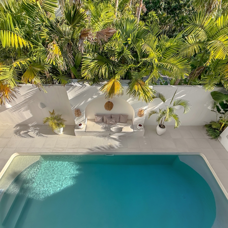 Outdoor pool area with white patio, built-in seating, and dense palm foliage overhead.