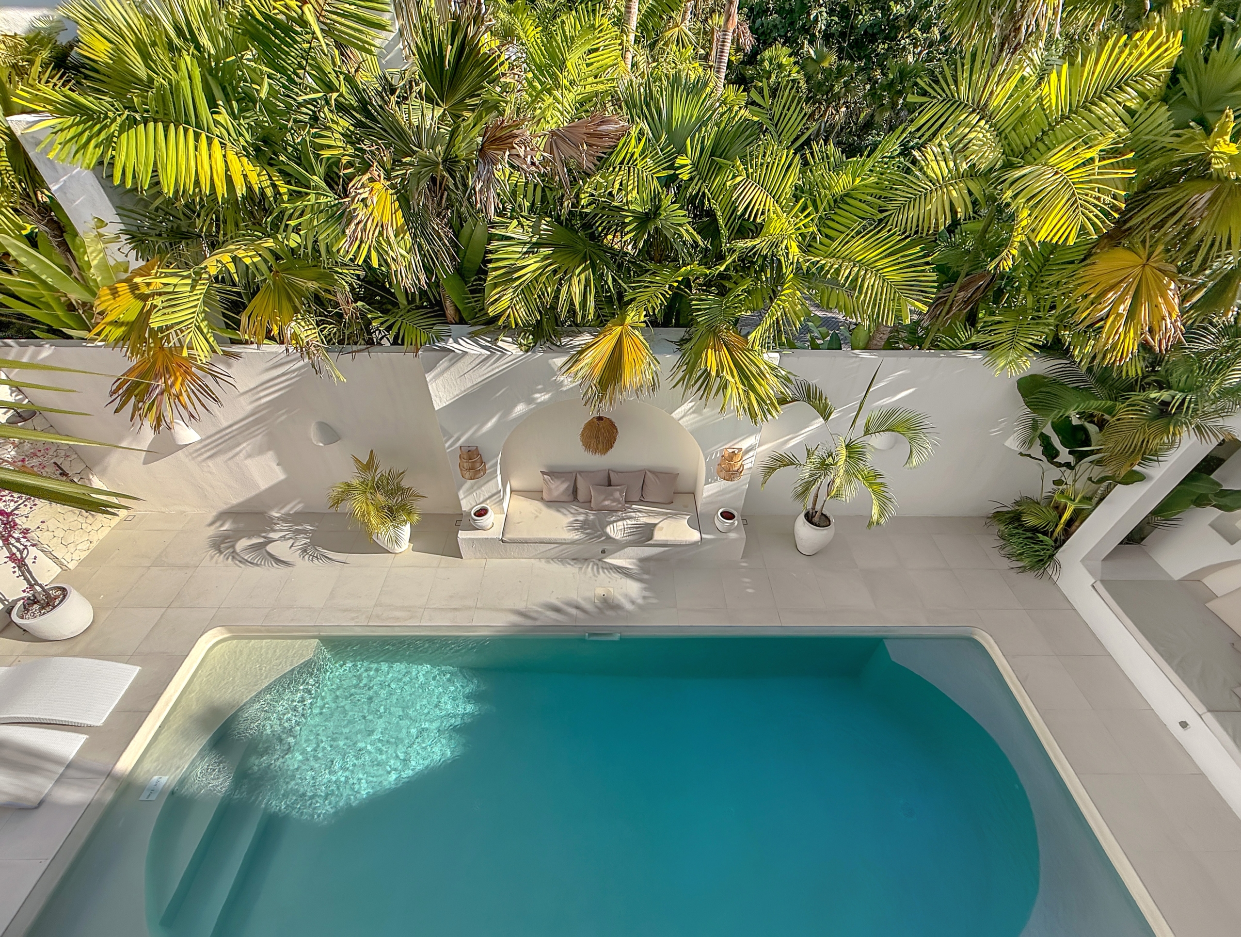 b6e4cfa1cc065a93862aaf9b04c53d1ca4d2e17c Outdoor pool area with white patio, built-in seating, and dense palm foliage overhead.